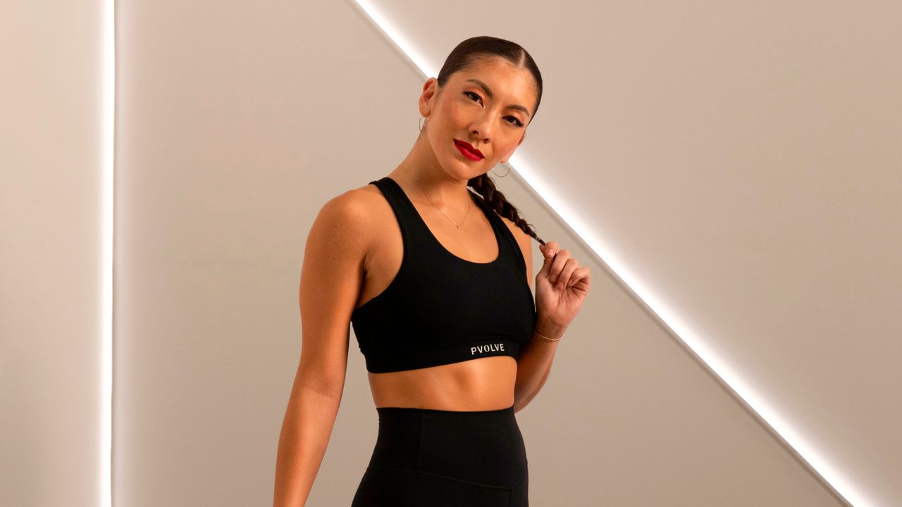 Woman in black sportswear looks at the camera. She is in a studio with strip lights behind her