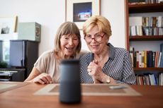 Two women look at a smart speaker excitedly. 