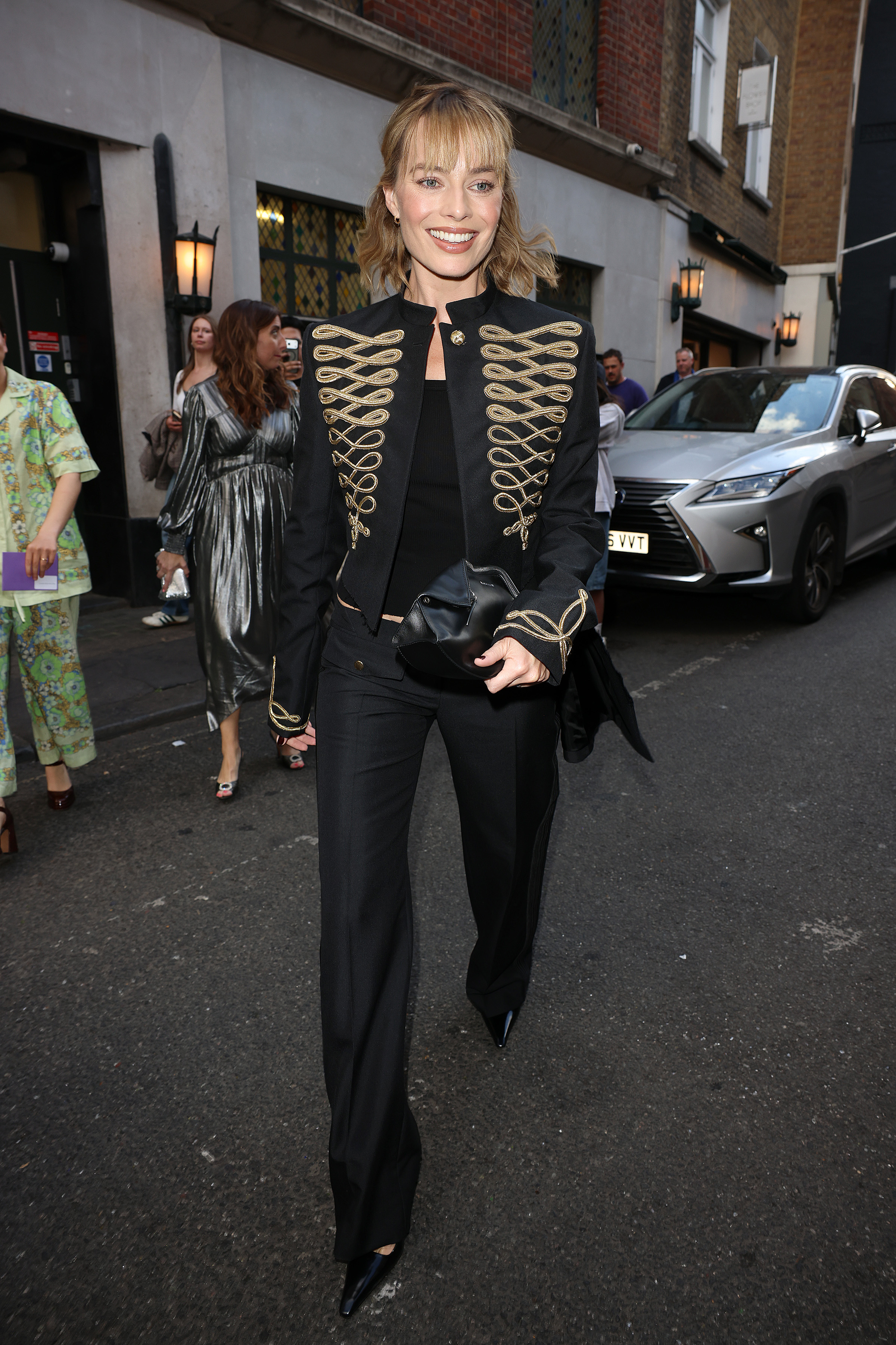 Margot Robbie steps outside in London wearing a military jacket with black low-rise trousers and pointed-toe heels.