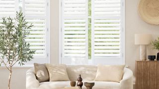 A bright, modern living room with white plantation shutters filtering soft daylight. A curved cream sofa with neutral cushions faces a wooden sideboard and potted olive tree, creating a calm, natural look.
