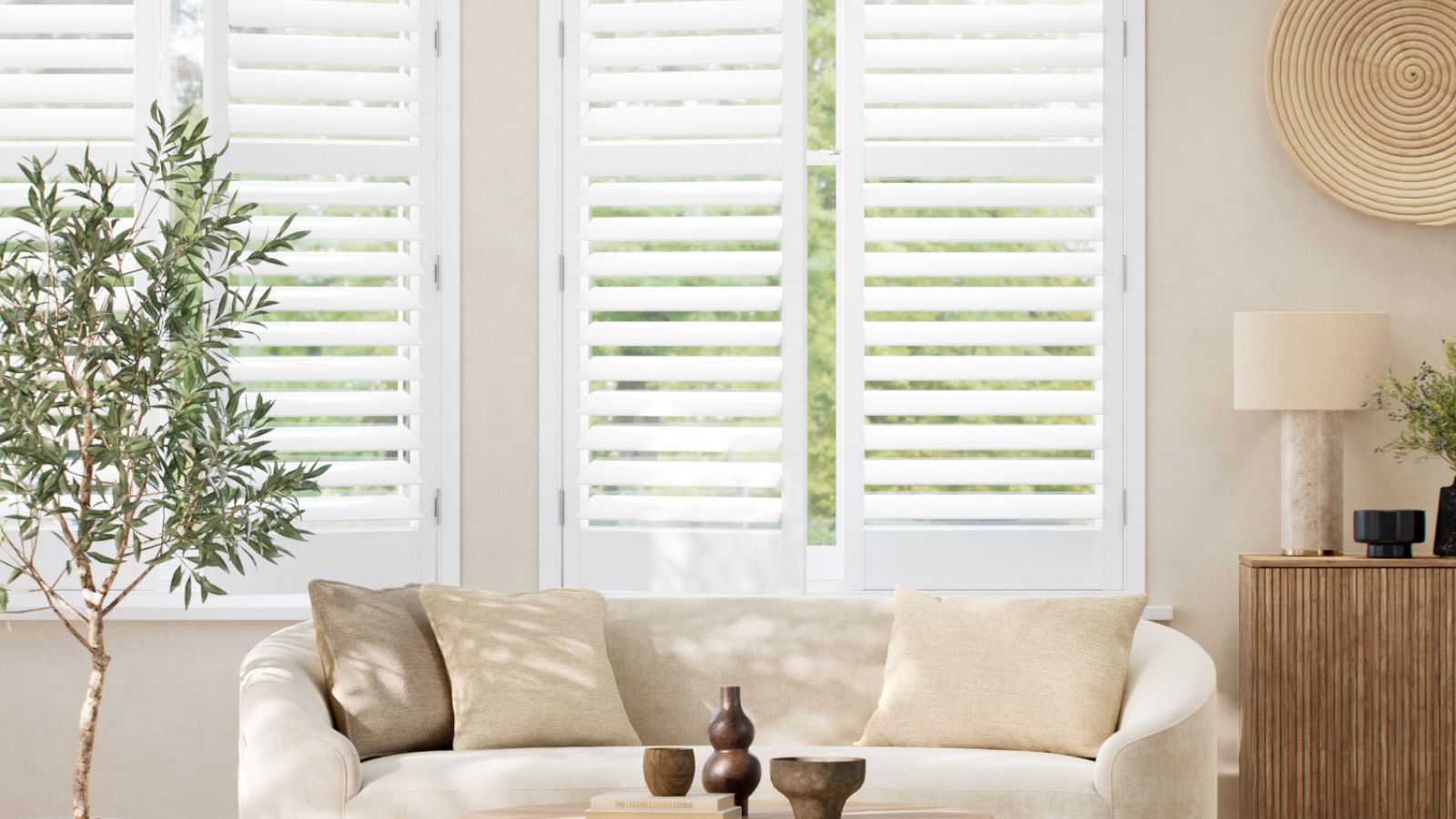 A bright, modern living room with white plantation shutters filtering soft daylight. A curved cream sofa with neutral cushions faces a wooden sideboard and potted olive tree, creating a calm, natural look.