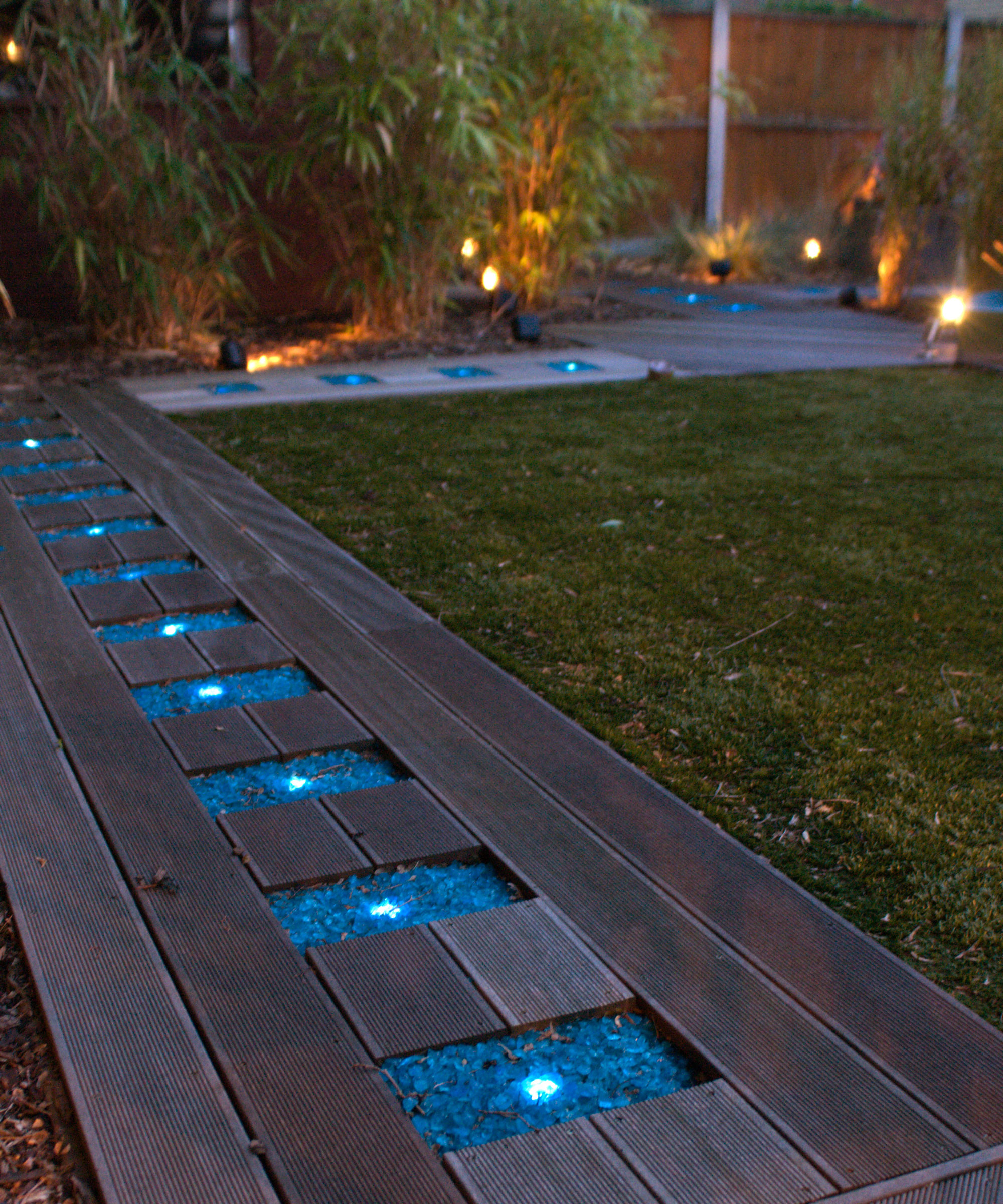 decking path with recessed LED lights set in gravel, bordering lawn, with lanterns and bamboo