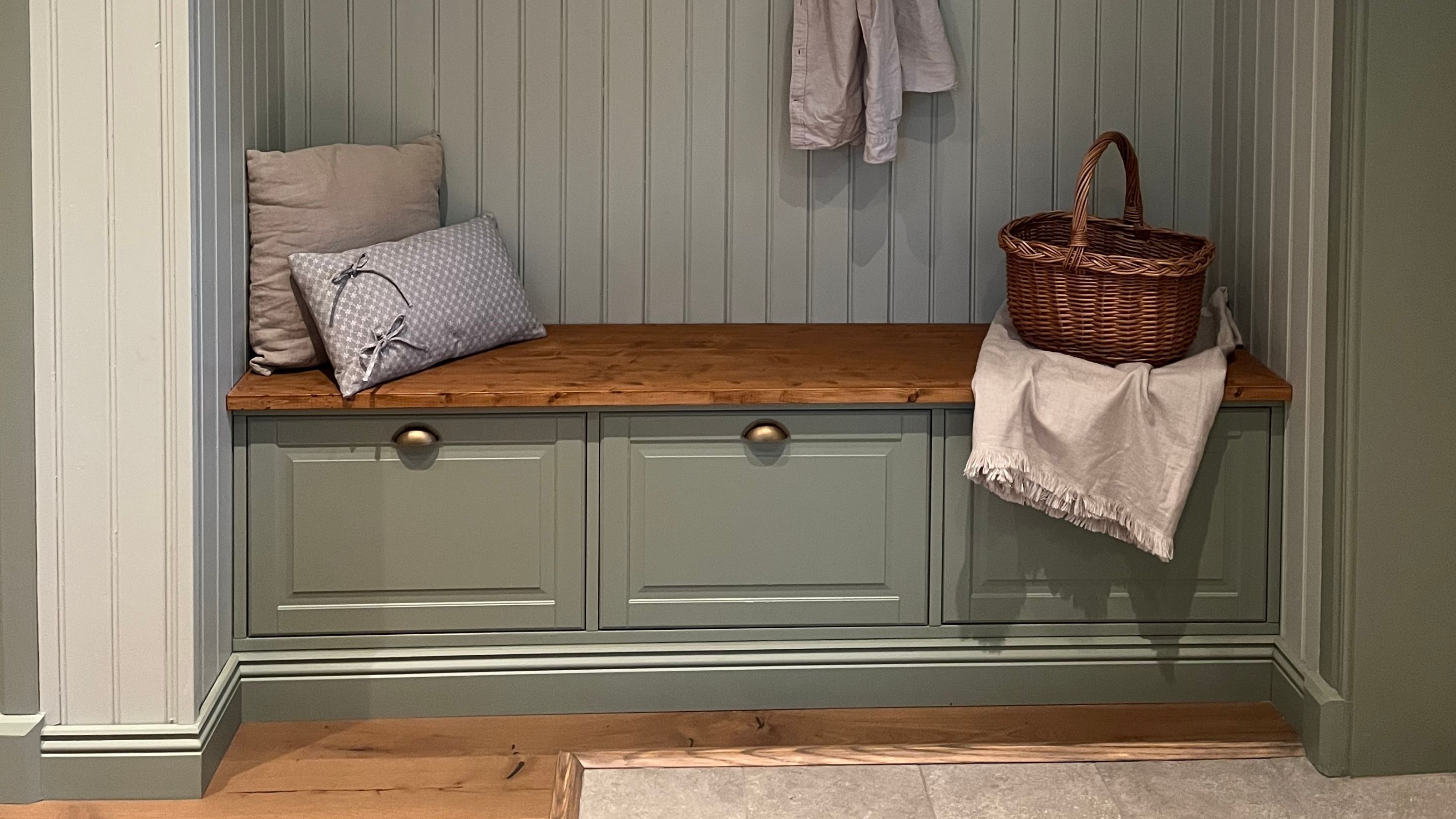 Built-in mudroom bench with sage green drawers, a warm wood top, decorative pillows, and a wicker basket against a matching beadboard wall