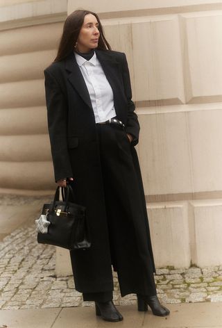 a photo showing what shoes to wear with wide-leg pants with a woman standing outside against a tan building wearing a long black coat layered over a white button-down shirt with black wide leg trousers and round toe ankle boots and a black Hermes bag