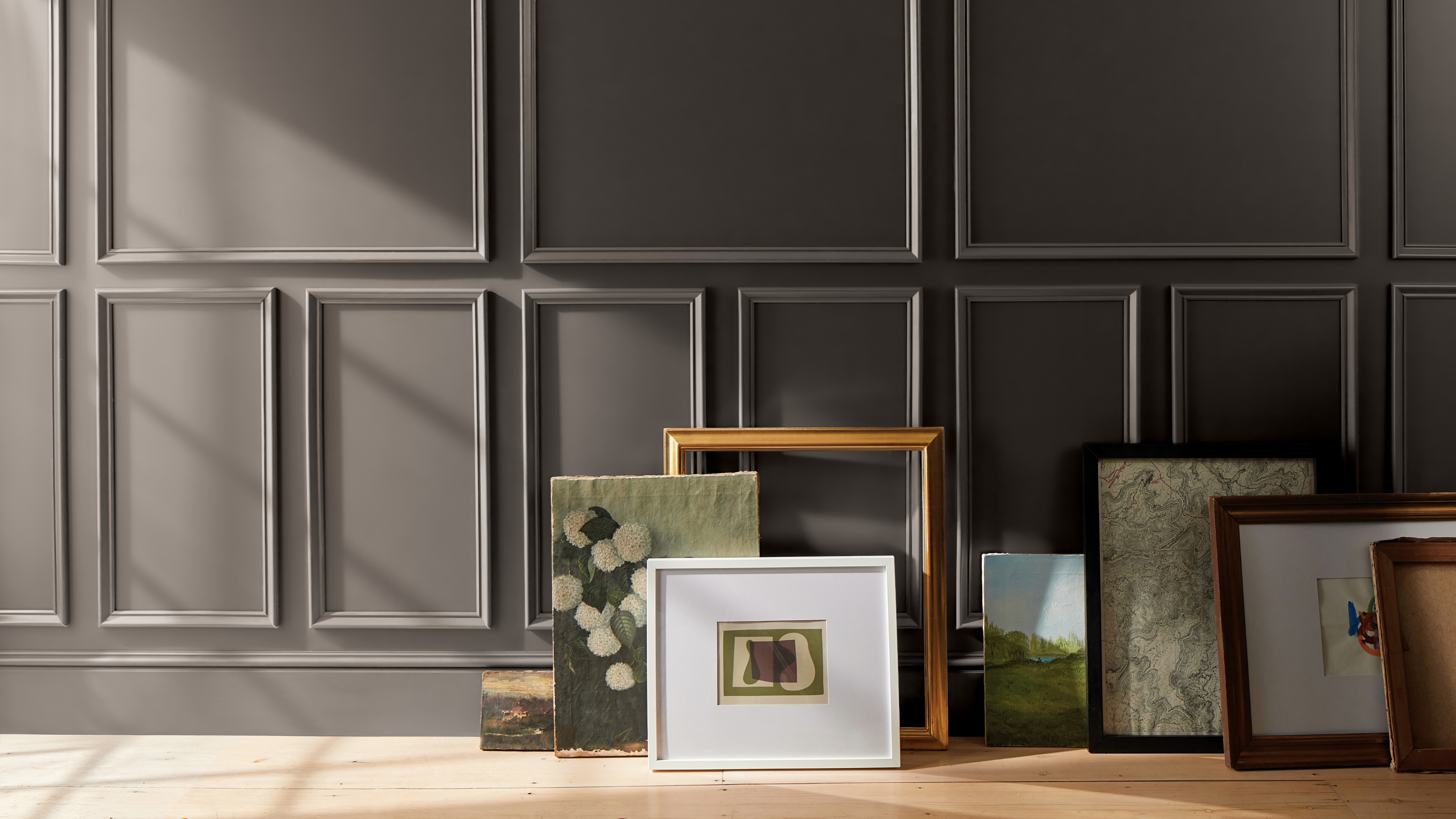 Image of a wall will crown molding that is painted in a dark purplish-gray wall color. There are lots of picture frames leaning against the wall.