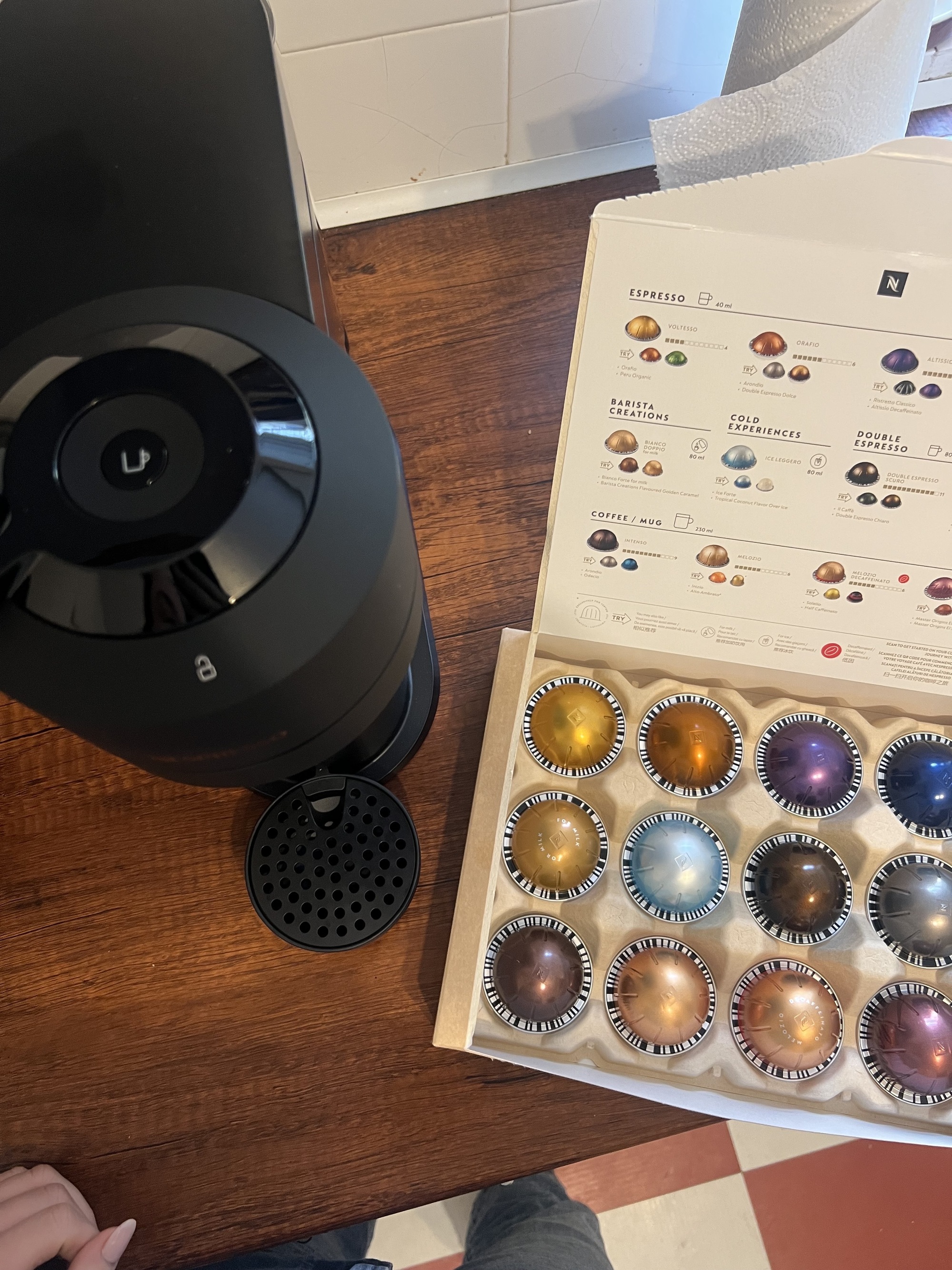 Image of a box of multicolored coffee pods on a wooden countertop next to a black Nespresso Vertuo Next machine.