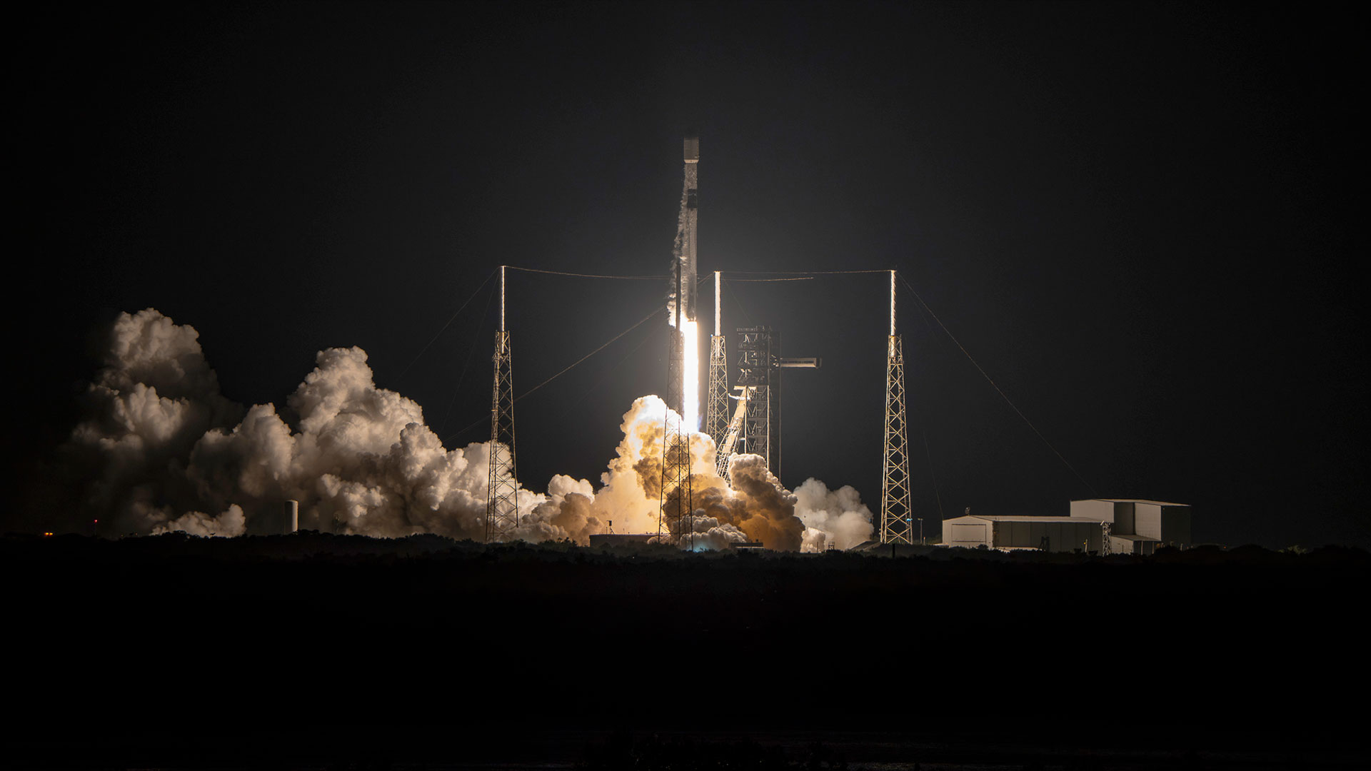 a white and black rocket lifts off into the night sky, its orange-white thrust lighting up its launch pad and billowing plume.