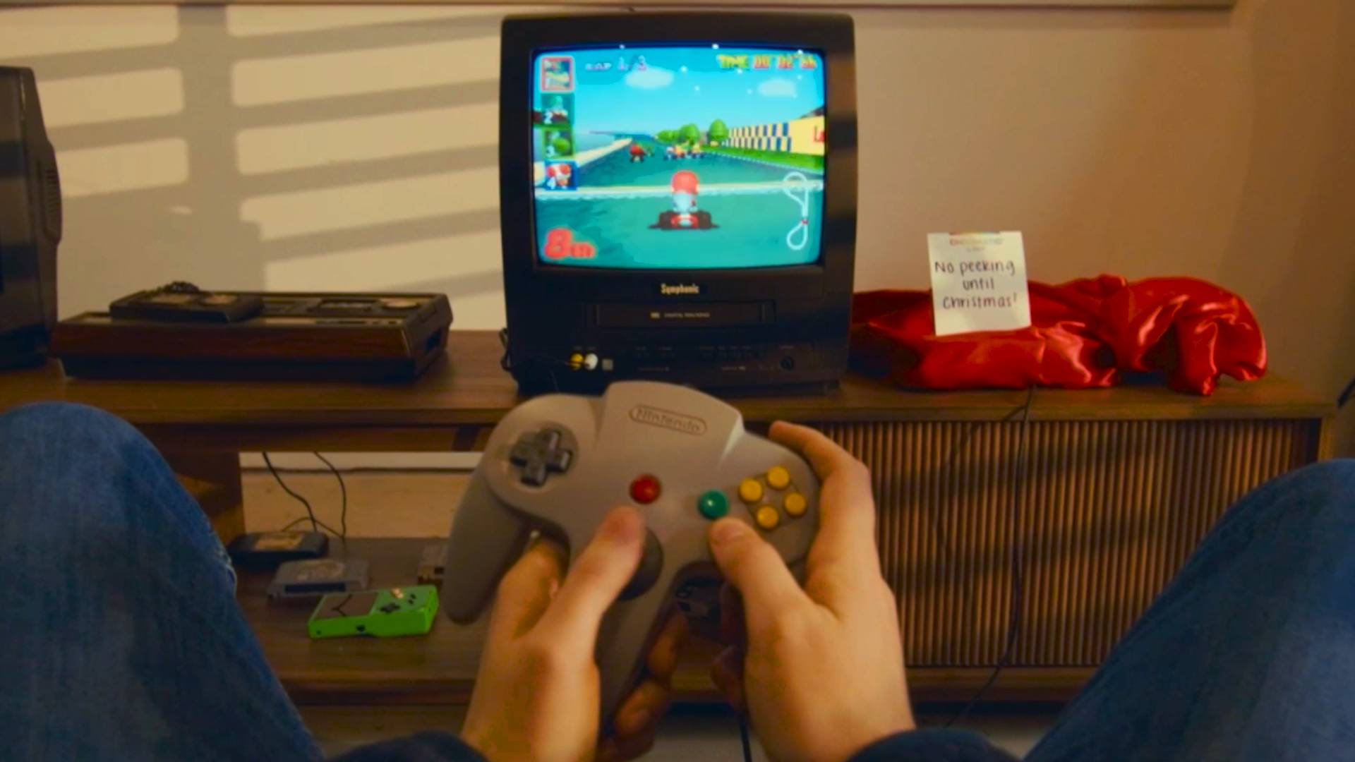 Player holding N64 controller in front of CRT with Mario Kart 64 on screen and console covered in red cloth next to sign that reads "no peeking until Christmas!"