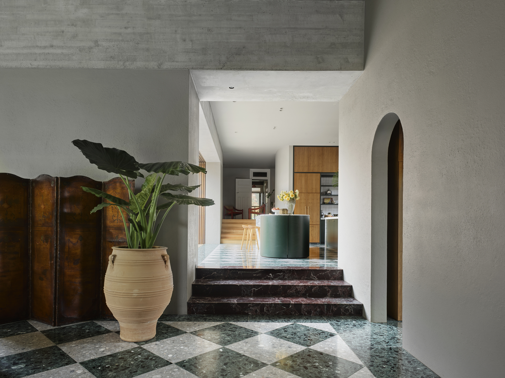 entryway with green marble checkerboard flooring with rendered walls, cement ceiling, large plant in terracotta pot and antique room divider screen, with red marble stairs leading up to green kitchen