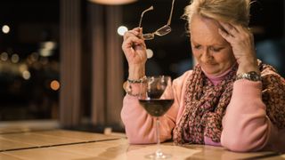 Woman sat at a table with her left hand on her forehead looking like she is in pain with a glass of red wine in the foreground