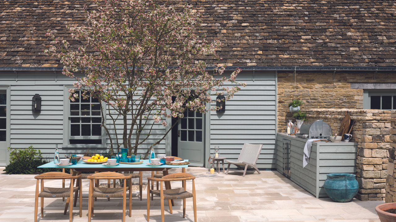 Outdoor kitchen area in garden with table and wooden chairs on patio area behind house with blue panels