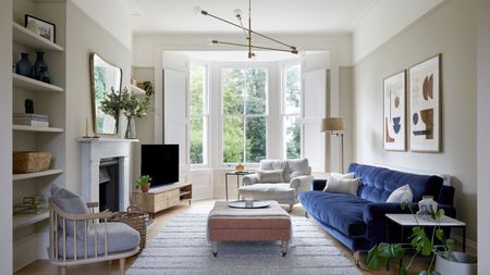 Living room with taupe walls, large bay window, grey rug at the centre, a pink ottoman on top, dark blue velvet sofa, and a large fireplace with a mirror above it