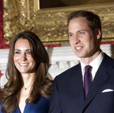 Kate Middleton wearing a blue dress and Prince William standing in front of a red wall and gold painting wearing a suit and tie