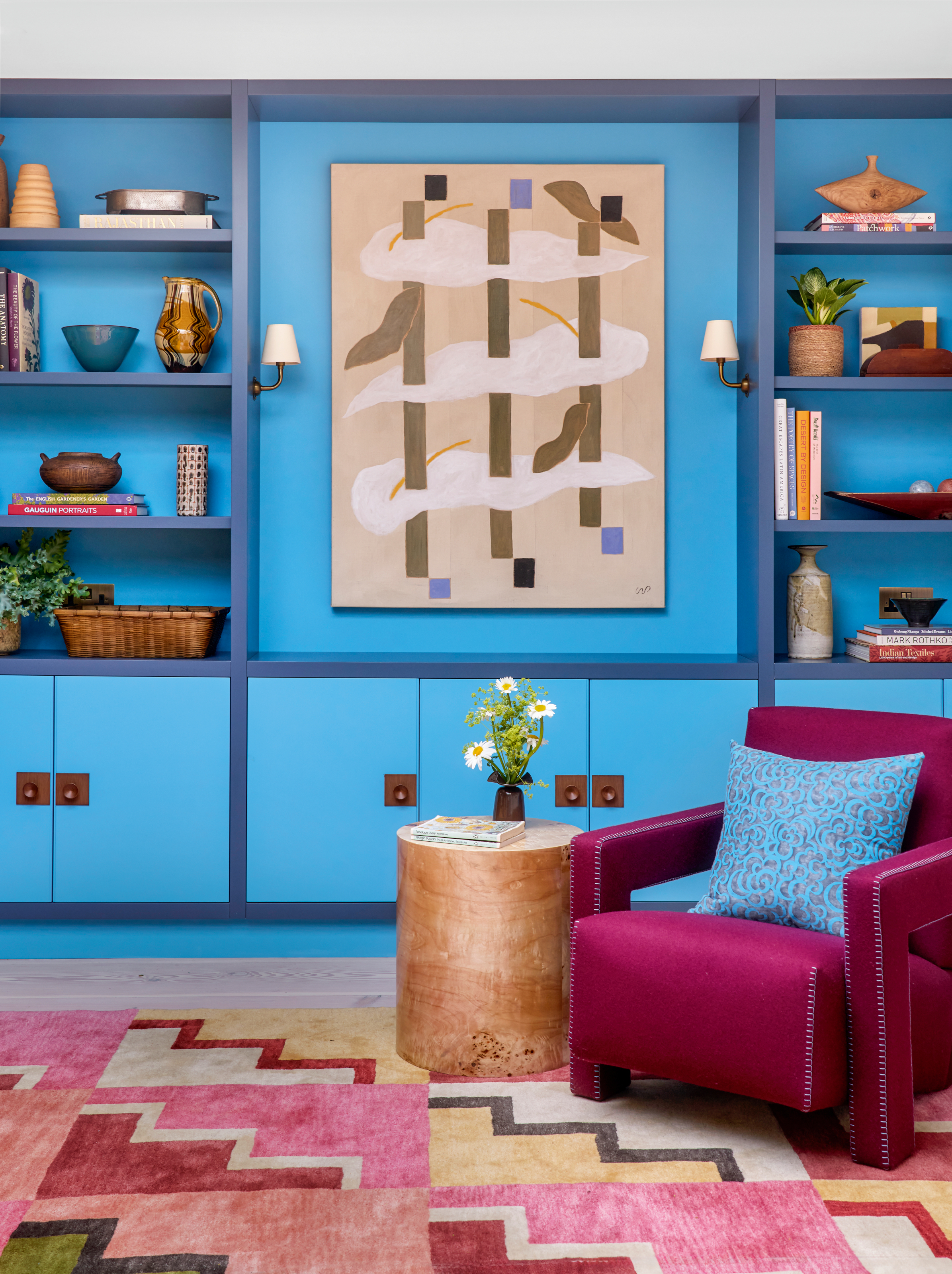 Corner of a living area with wall of joinery in bright blue, a red and pink geometric rug, a bright pink armchair and burl wood side table