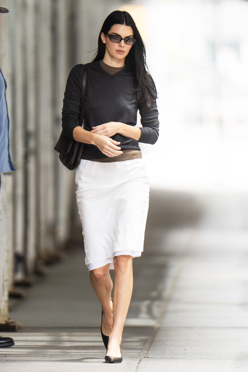 Kendall Jenner wearing a gray V-neck sweater over a brown T-shirt with a white knee-length skirt and black kitten-heel pumps in NYC