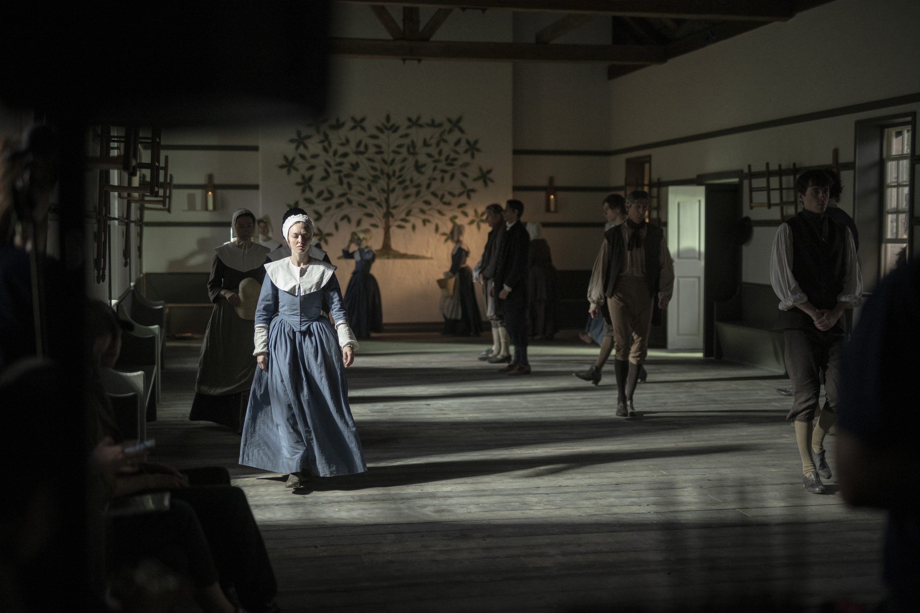 A small group of people wearing 18th-century clothing walk around a meeting room with a tree decoration on the back wall and Shaker chairs on the right wall. One woman (Amanda Seyfried as Ann Lee) stands in a ray of sunlight, while the rest are in shadow, in a still from 'The Testament of Ann Lee.'