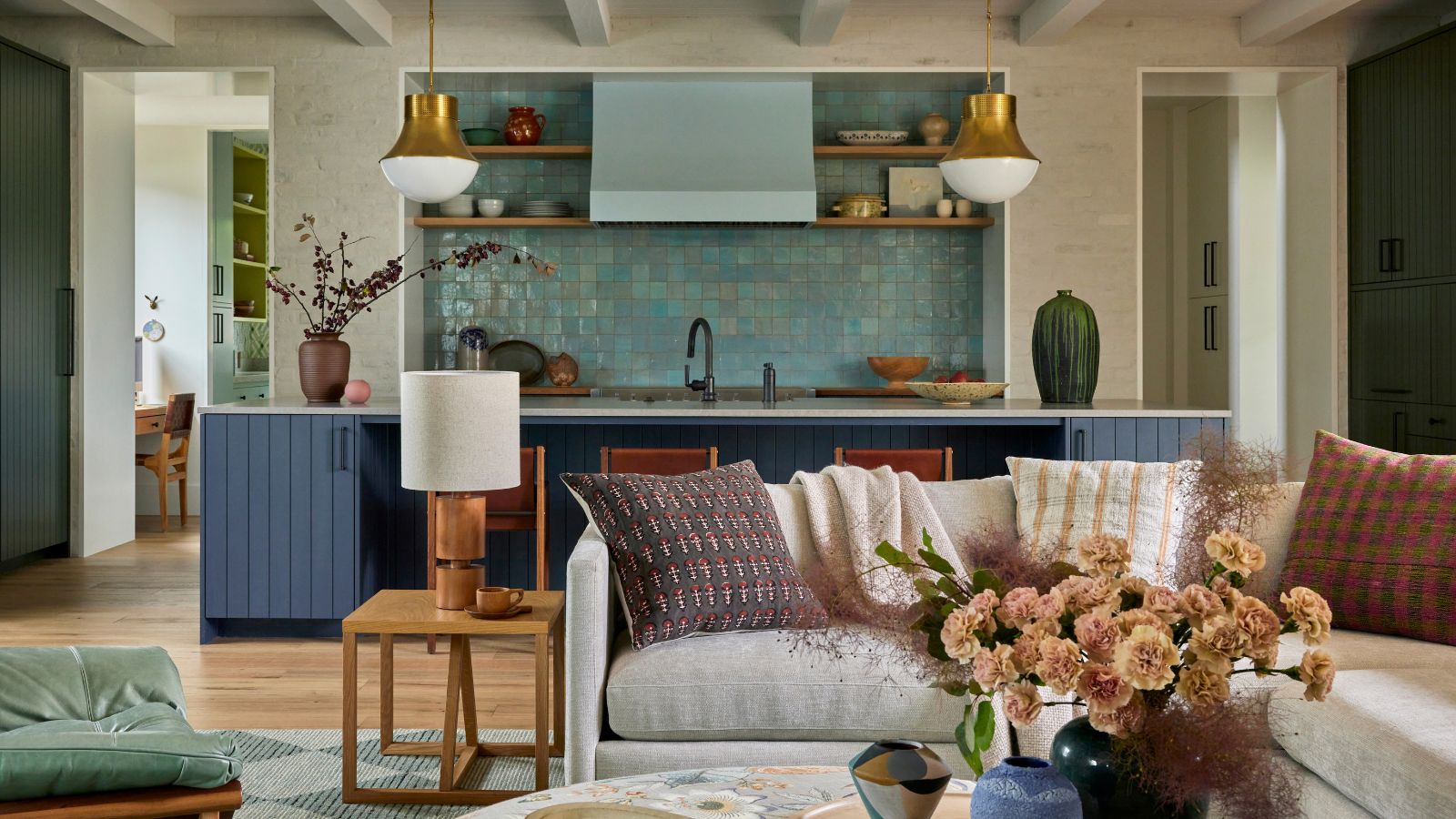A modern, open-plan living room and kitchen. A teal blue tiled backsplash, dark blue cabinets, with rustic decor and two brass pendant lights. A gray sectional with patterned scatter cushions, a round coffee table with a tray and vase of flowers, and a wood side table with a table lamp.