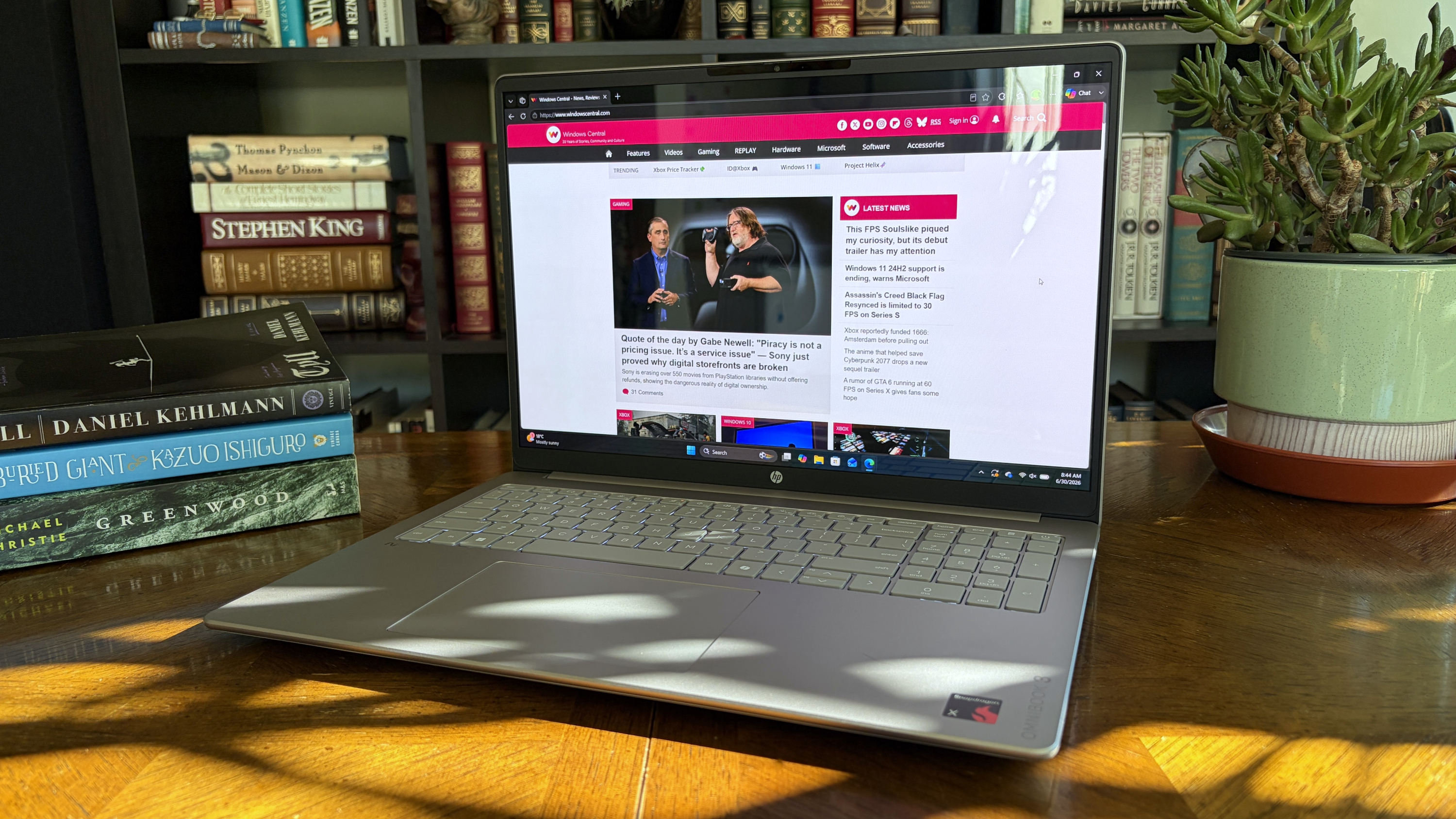 A modern silver OmniBook 3 laptop on a wooden table displays a gaming news website, surrounded by stacked colored books and a potted succulent.