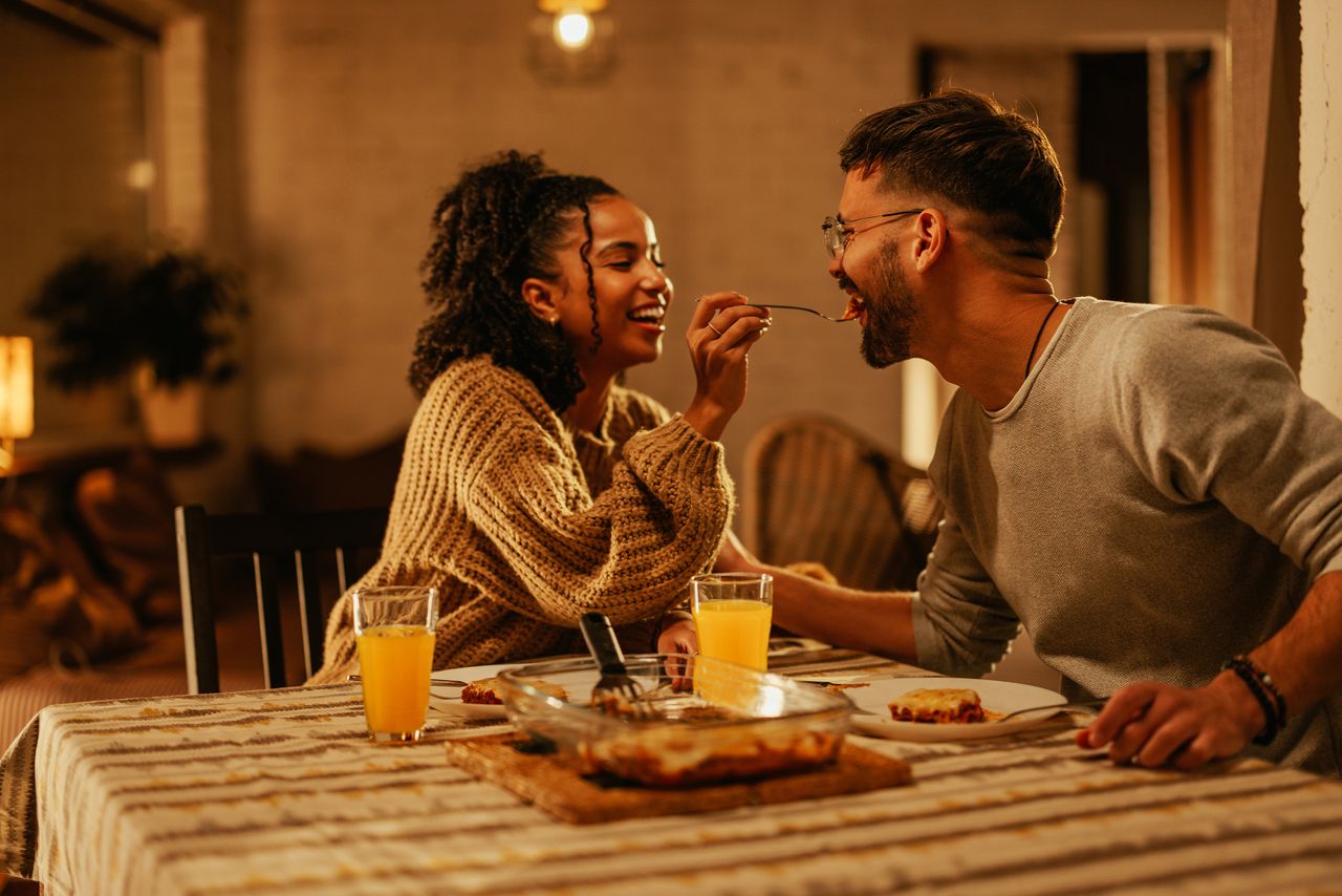 Happy couple eating a romantic dinner at home, with the woman spooning a mouthful of food into the man&#039;s mouth.