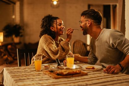 Happy couple eating a romantic dinner at home, with the woman spooning a mouthful of food into the man's mouth.