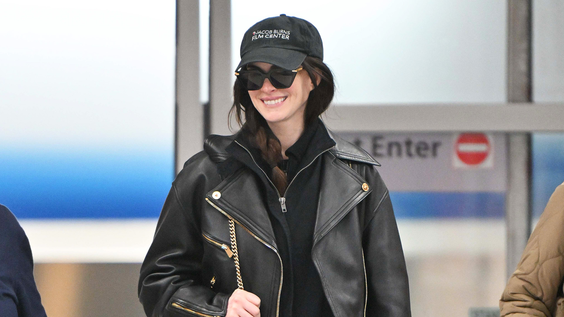 Anne Hathaway at the airport in NYC wearing a black leather jacket, half-zip pullover, black pants, black chunky shoes, a black quilted bag, and baseball cap