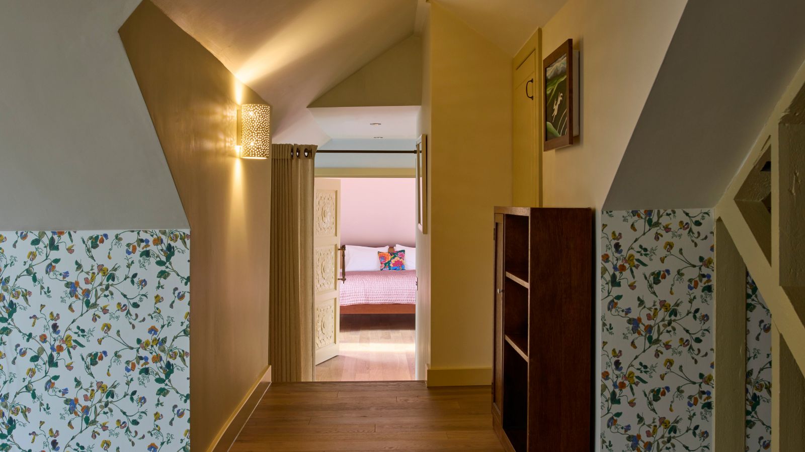 A narrow loft hallway with sloped ceilings and warm yellow walls above floral wallpaper. Wooden flooring leads to an open carved door, revealing a pink bedroom beyond. A wall sconce glows softly, and a small wooden shelving unit sits to the right.