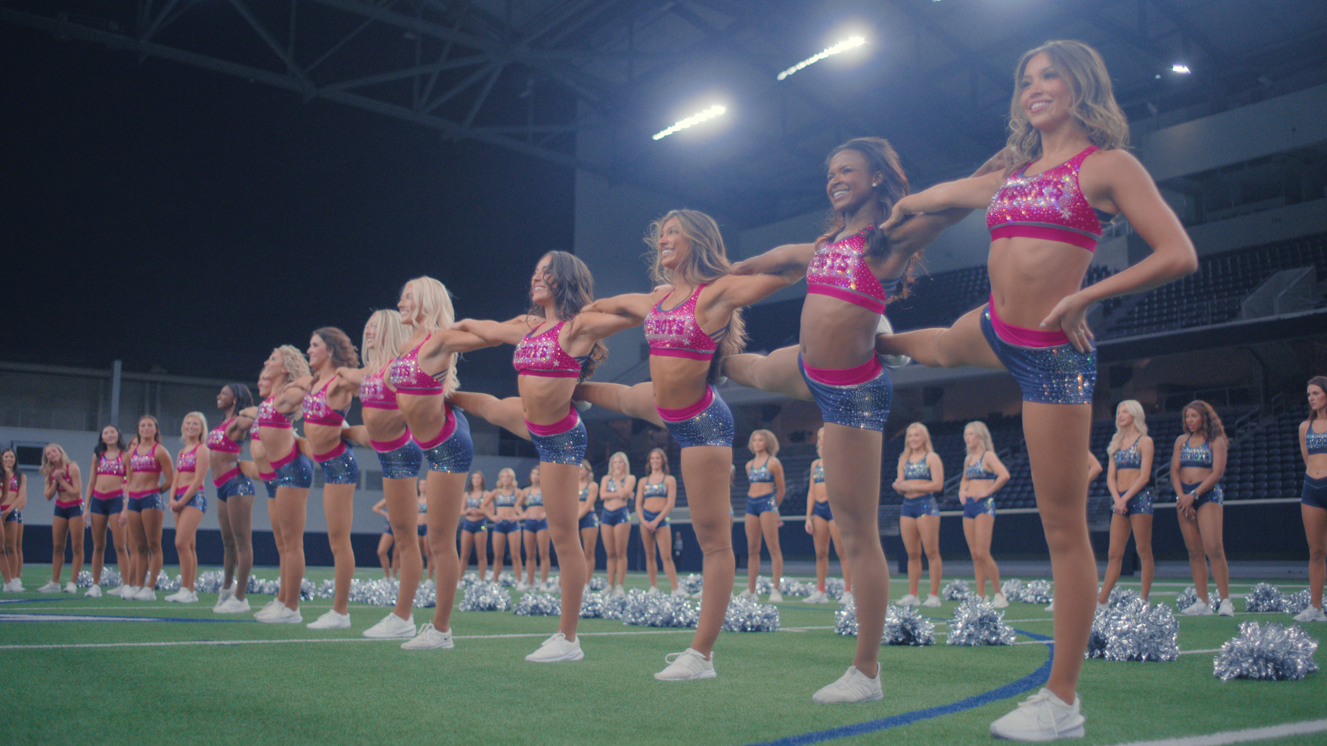 A line of cheerleaders stand on one leg while other dancers stand around them in a practice facility, in 'America’s Sweethearts: Dallas Cowboys Cheerleaders.'