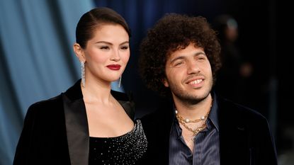 Image of Selena Gomez in a black blazer and diamond earrings standing next to Benny Blanco, who is wearing a black blazer and blue satin shirt