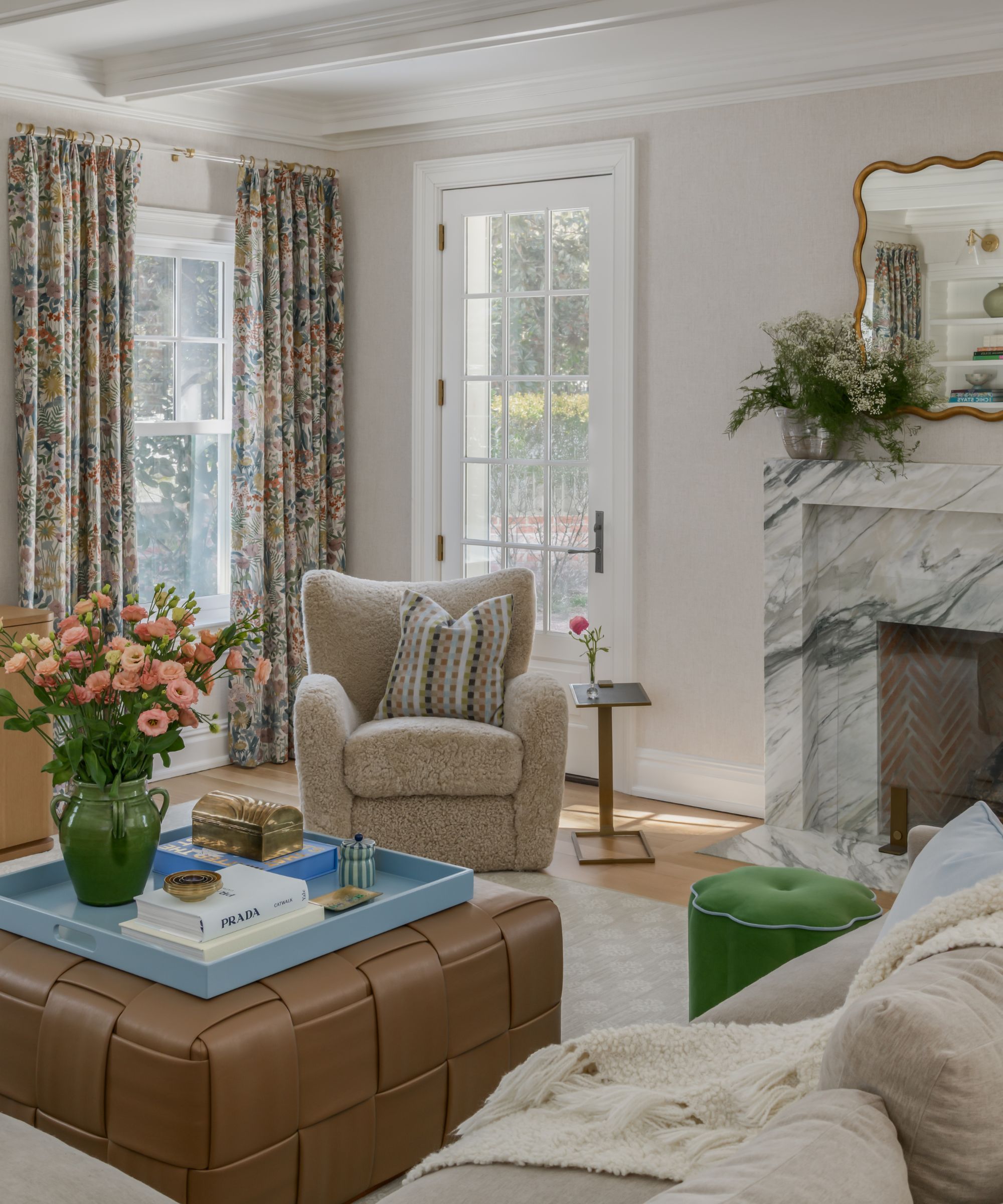 A transitional living room with off-white walls, a marble fireplace, A French door, and a large window with floral full-length curtains, A beige armchair, a sectional, and a brown ottoman with a blue tray and vase of pink flowers.