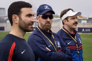 jason sudeikis as coach ted lasso standing next to the assistant coach and a soccer player on a field in the tv show ted lasso