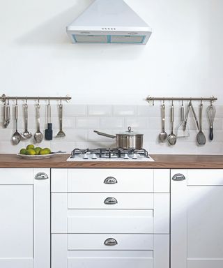 White kitchen units with a wood worktop, hob and extractor hood, and utensils hanging from rails.