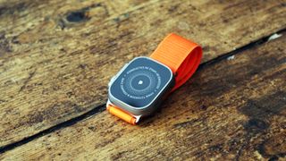 En Apple Watch Ultra med ett orange textilband ligger utomhus på träplankor.