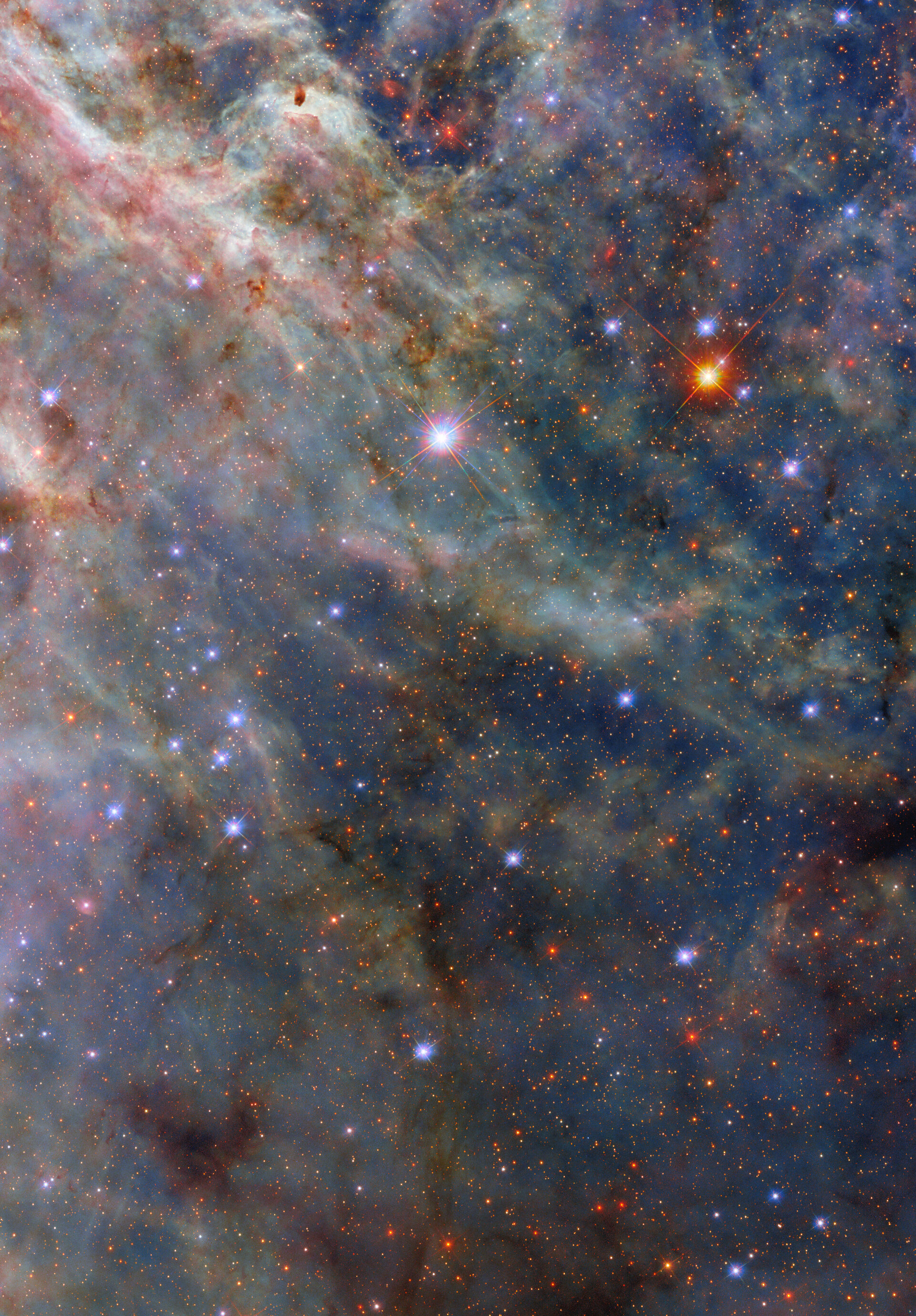 A nebula. The top-left is dense with layers of fluffy pink and greenish clouds. Long strands of green clouds stretch out from here; a faint layer of translucent blue dust combines with them to create a three-dimensional scene. A sparse network of dark dust clouds in the foreground adds reddish-black patches atop the nebula. Blue-white and orange stars, from our galaxy and beyond, are spread amongst the clouds.