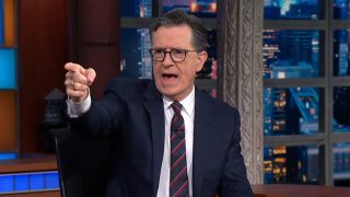 Stephen Colbert points off stage during one of the final episodes of The Late show. He is wearing a suit and tie. 