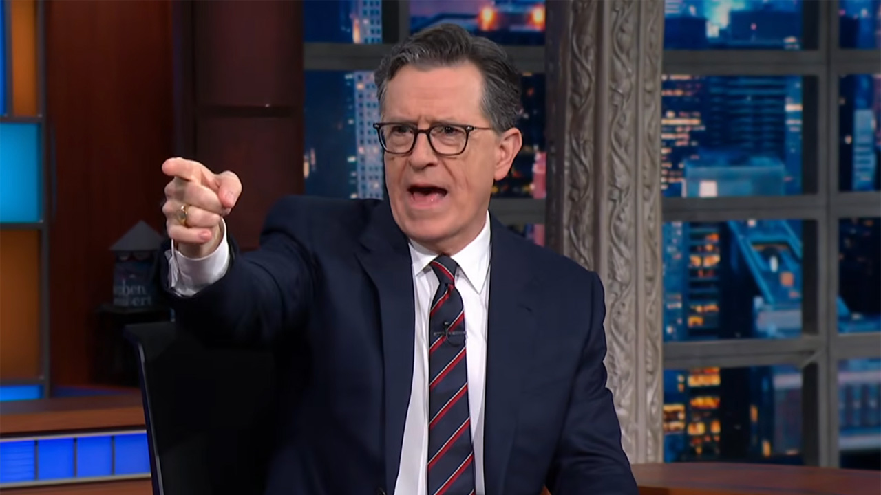 Stephen Colbert points off stage during one of the final episodes of The Late show. He is wearing a suit and tie. 