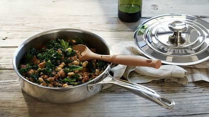 A stainless steel pan filled with broccoli and chickpeas, on a wooden table with a matching lid beside it on a dish towel. A wooden spoon inside the pot.