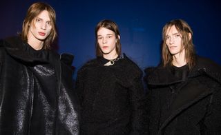 Three male models wearing looks from the Rick Owens collection. Two models are wearing black tops and black coats in different styles. And one model is wearing a black top with a black fluffy style piece over the top and has a chain and light coloured strap by the neck area. They are standing in front of a blue wall