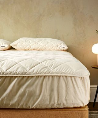 Woolroom mattress wrapped in a mattress protector on a bed base against a beige wall.