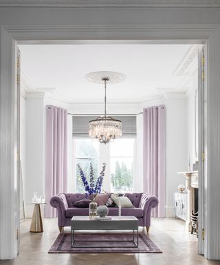 A white and lilac living room with statement chandelier light fixture and long light purple lavender curtains