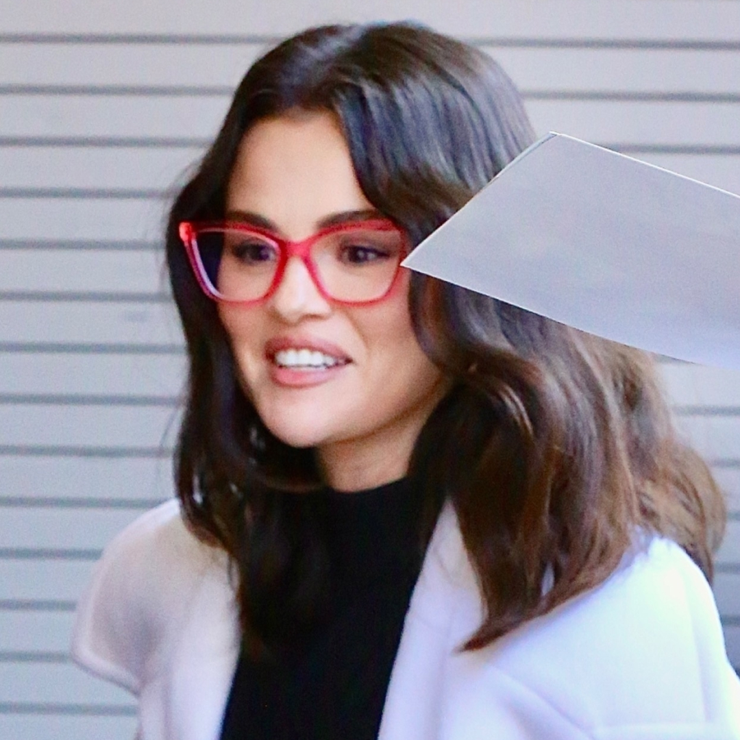 Selena Gomez wearing heeled flip-flops, a black dress, a white coat, and red glasses