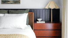 A white bedroom with black wall panelling, rattan bedframe with white bedding and green cushions, a red wood chest of drawers with white fluted lamp, small white bowl, and framed artwork above the bed