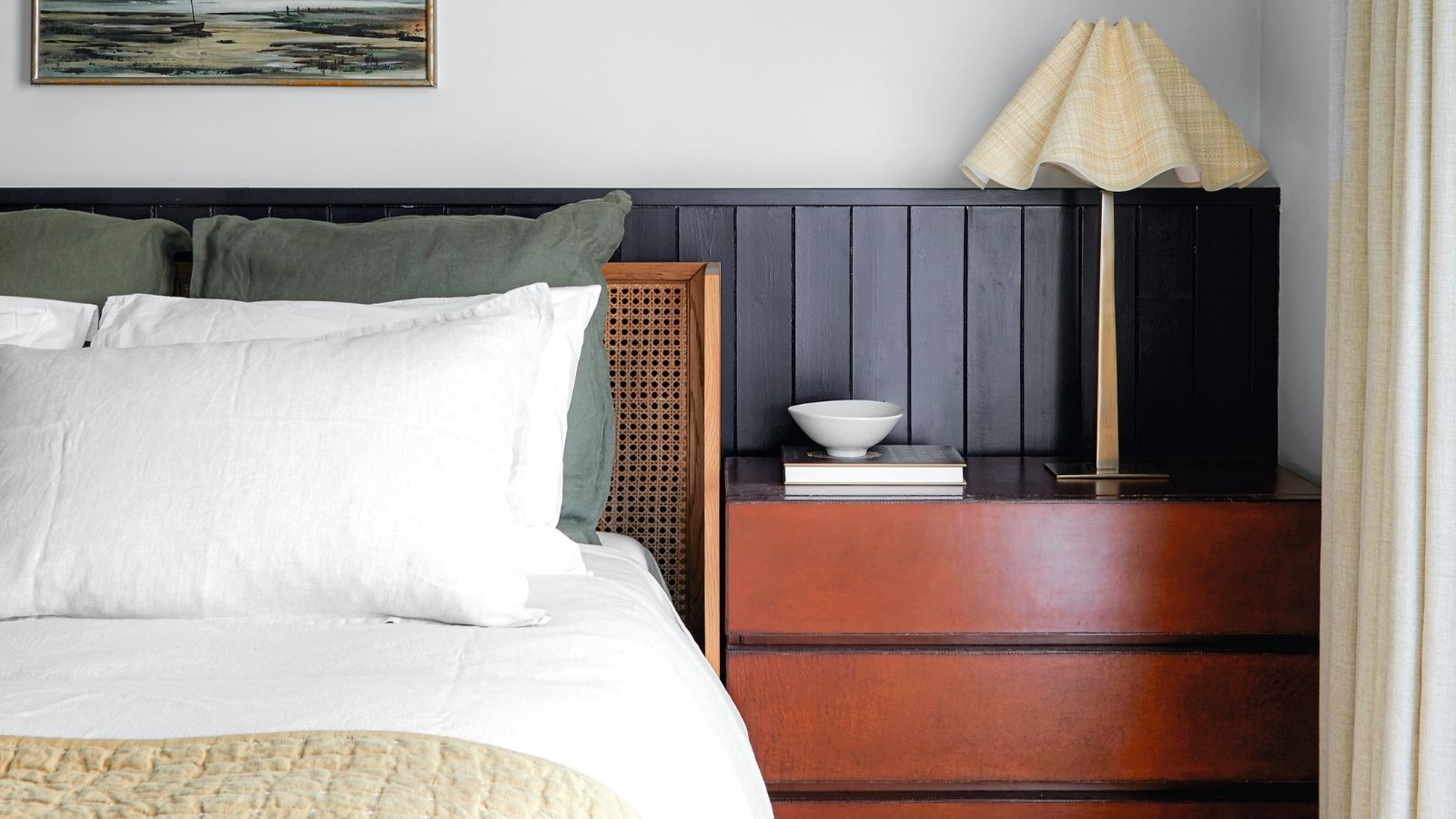 A white bedroom with black wall panelling, rattan bedframe with white bedding and green cushions, a red wood chest of drawers with white fluted lamp, small white bowl, and framed artwork above the bed