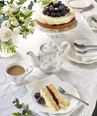 A white and gray tea pot on a white table cloth with a cherry cream cake