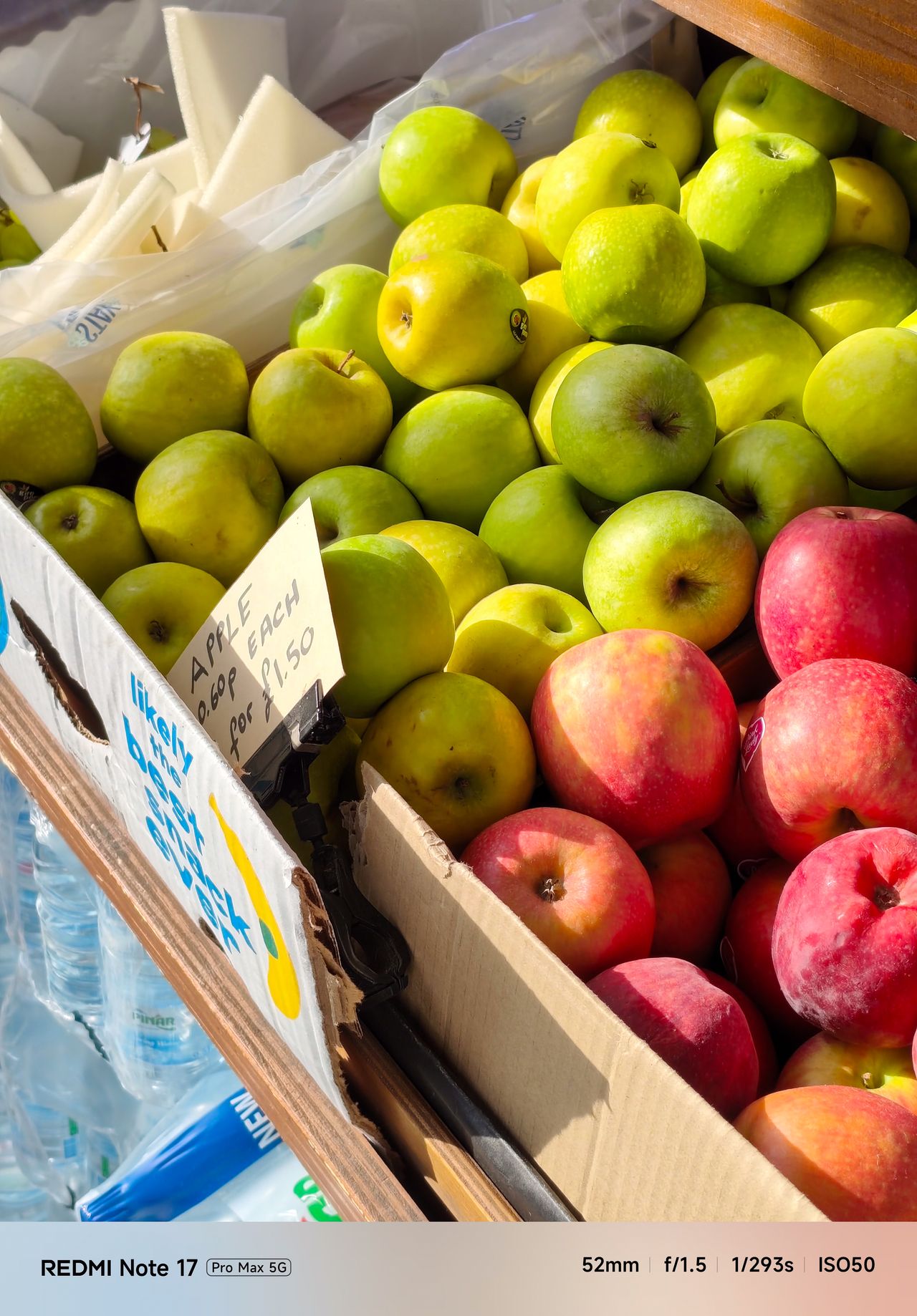 Red and green apples arranged in boxes at a market stall, photographed in bright sunlight.