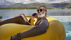 A retired man sips a tropical drink while lounging in a pool on an inflatable ring.