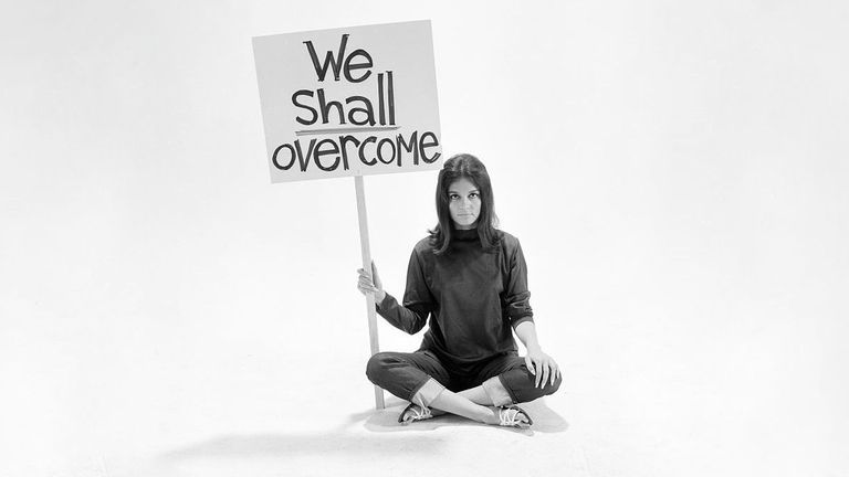 A woman in 1960s clothes holding a placard that says "we shall overcome"