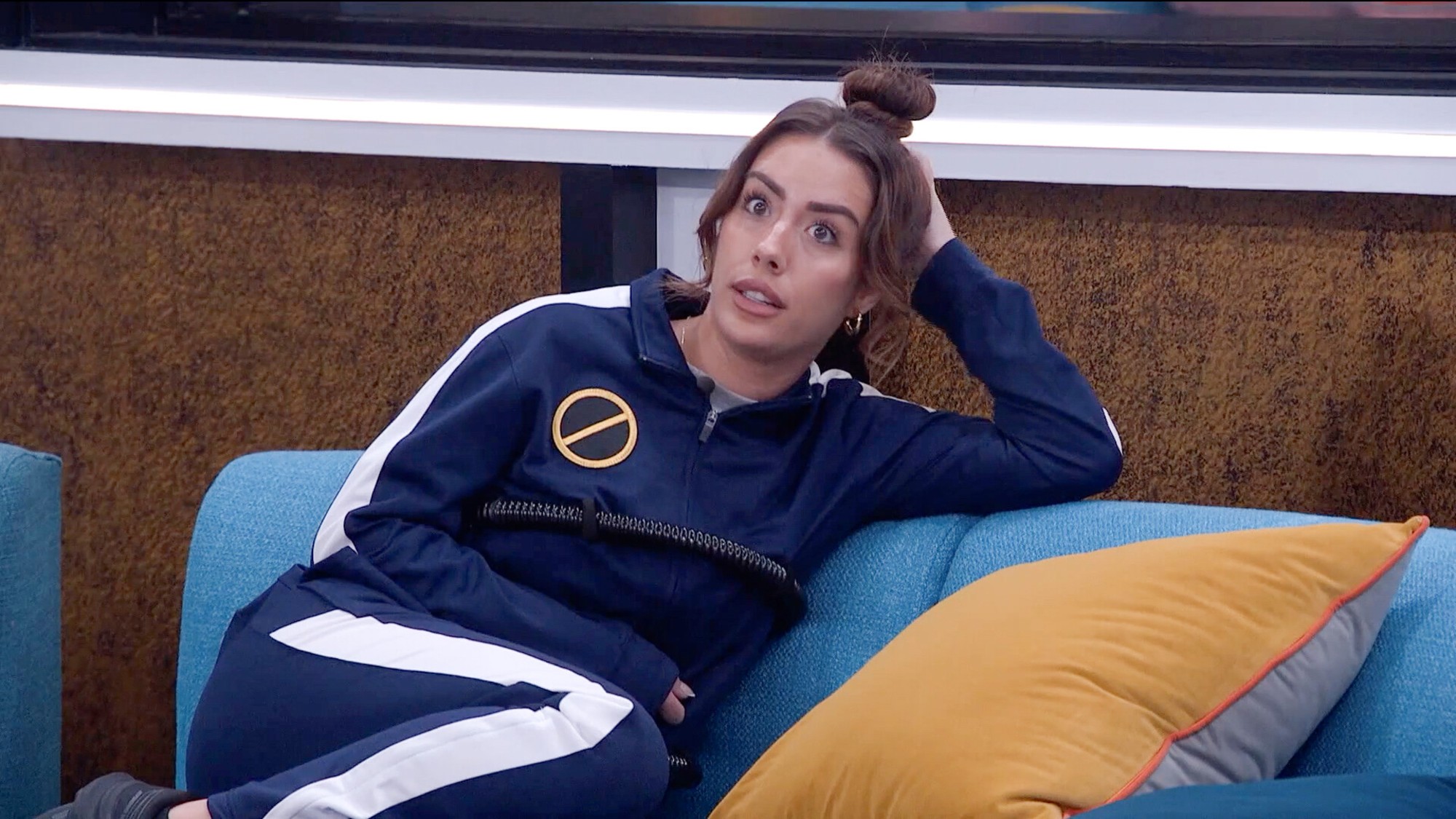 Dee watching the competition unfold while wearing a veto necklace and a blue track suit. Her hair is in a tight bun on top. 