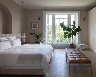 Large master bedroom leather daybed at foot of bed, traditional white windows, arched alcove, fitted bespoke storage, large plant on floor, wooden parquet flooring