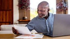 An older man smiles as he looks at paperwork while working on his laptop at the kitchen table.