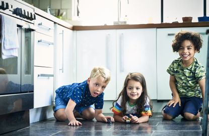 Three smiling children - two boys and a girl - playing with toy cars on a kitchen floor